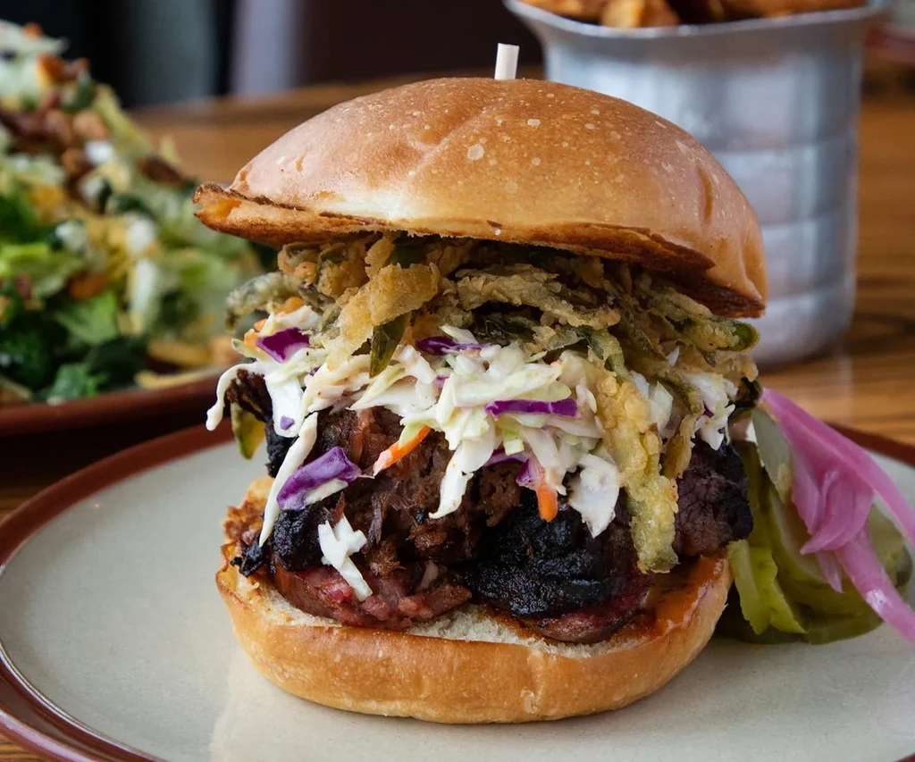 Stacked burger with coleslaw, pickles, and pickled onions on a plate.