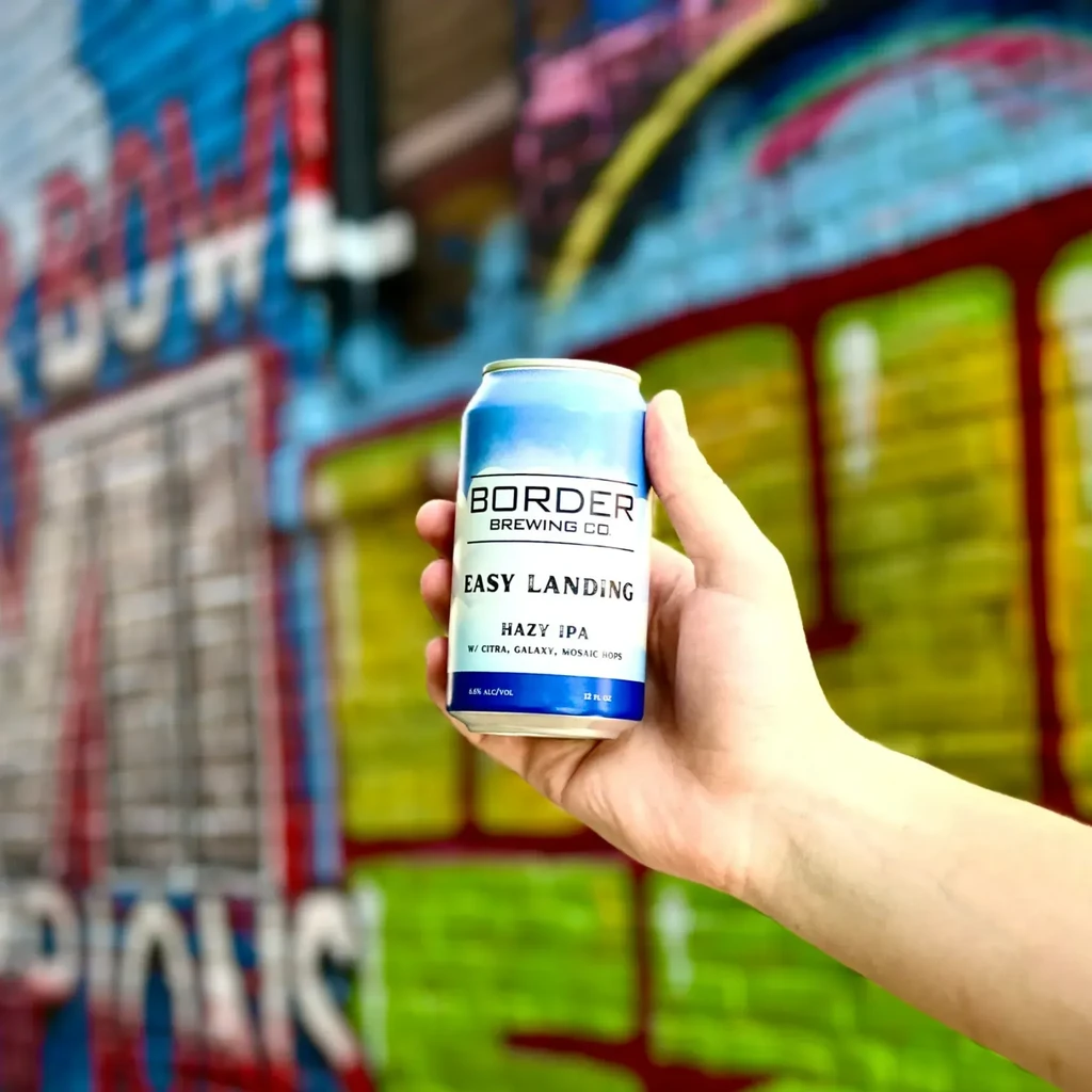 Hand holding a Border Brewing Co. Hazy IPA can against blurred graffiti.