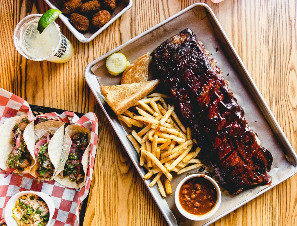 Overhead flat-lay: Ribs, fries, tacos, hushpuppies, corn salad, and a margarita.