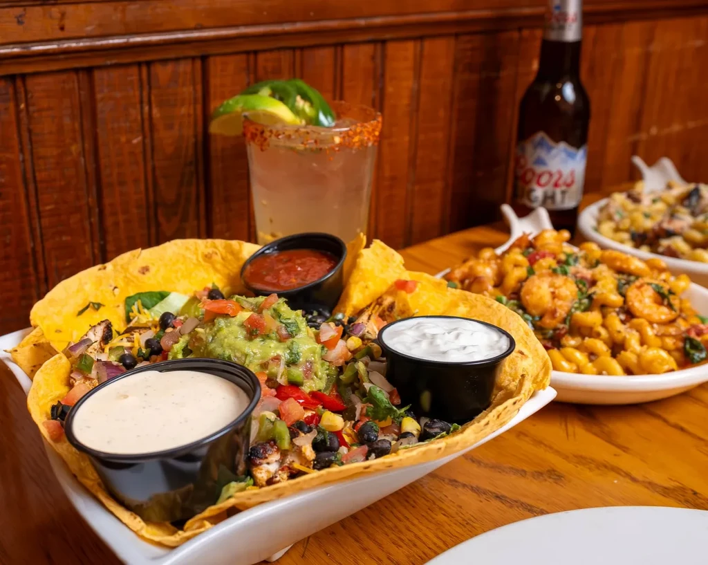 Loaded tortilla bowl salad, shrimp pasta, and drinks on a wood table.