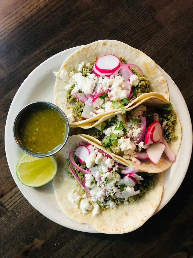 Overhead: Three tacos with green filling, cheese, radish, salsa, lime.