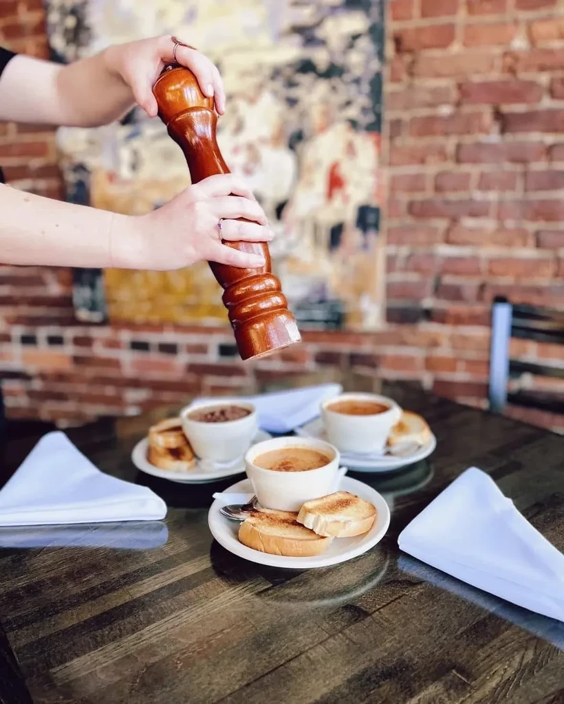 Hands grinding a pepper mill over soup bowls on a restaurant table.