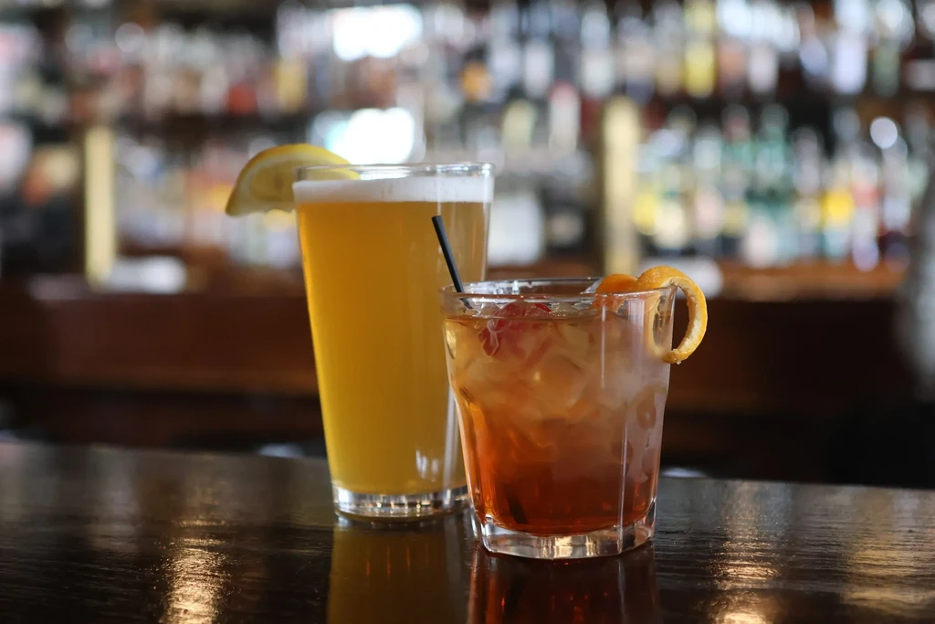 Two drinks: beer with lemon and a dark cocktail, on a bar.