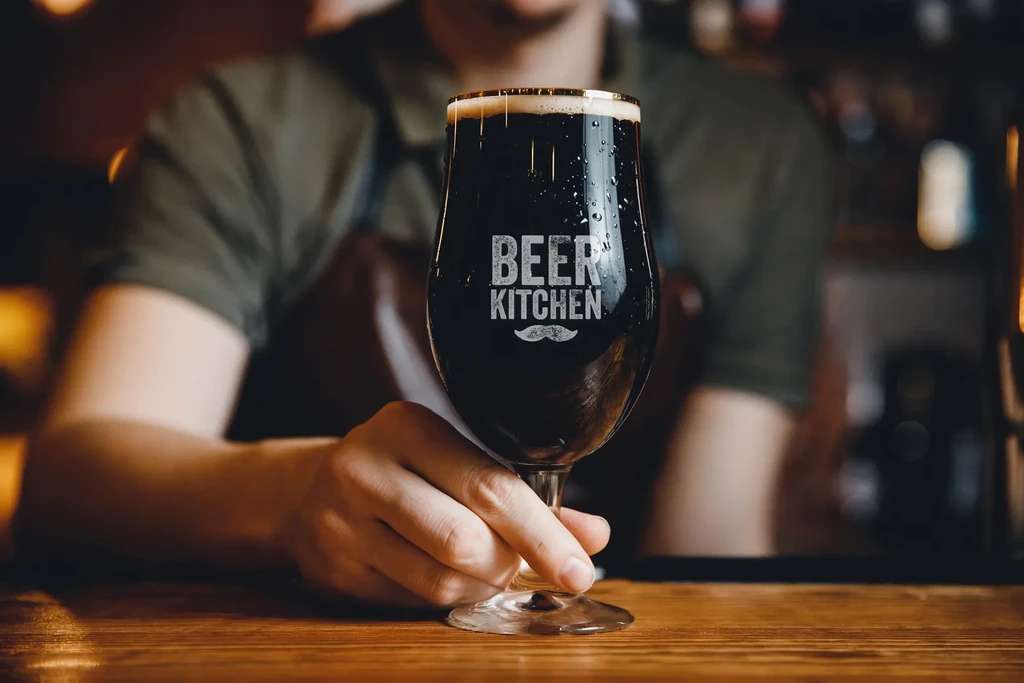 Close-up: hand holding a dark beer glass with condensation and 'BEER KITCHEN'.