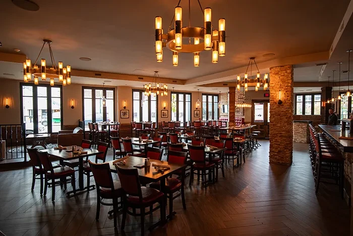 Warmly lit empty restaurant dining area, bar, chandeliers, stone pillar.