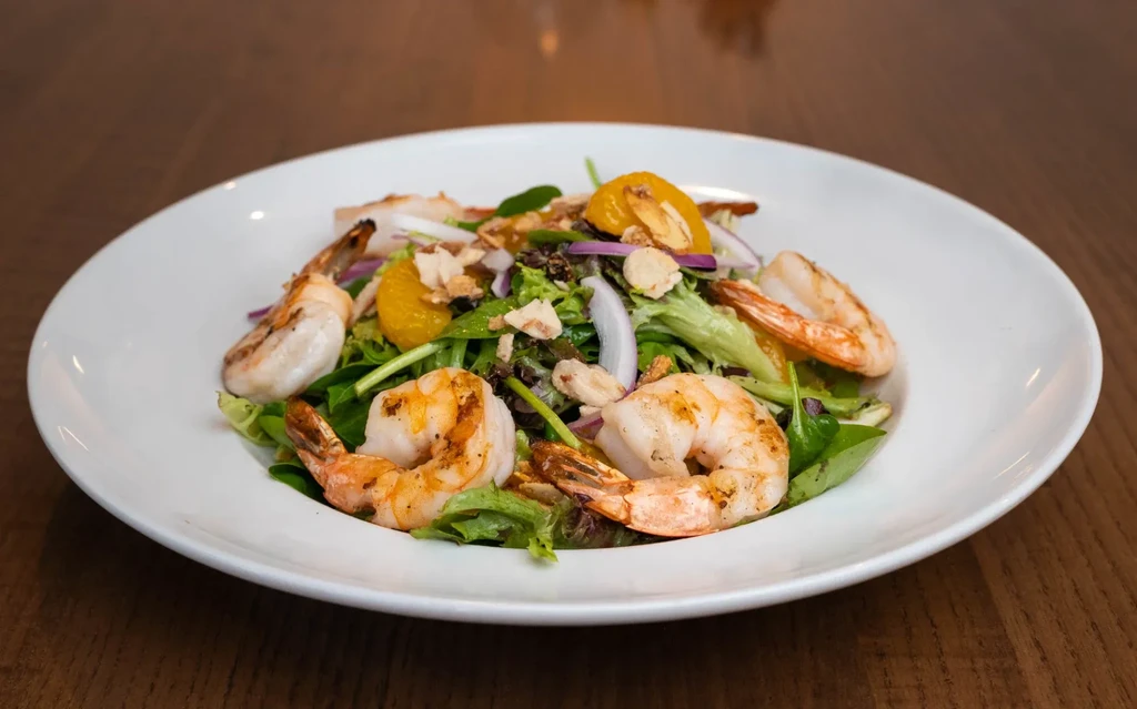 Grilled shrimp salad with greens, citrus, and nuts in a white bowl.