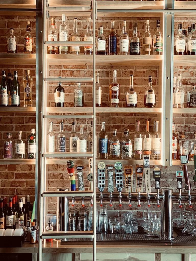 Stocked bar, bottles on shelves, beer taps, glasses, brick wall.