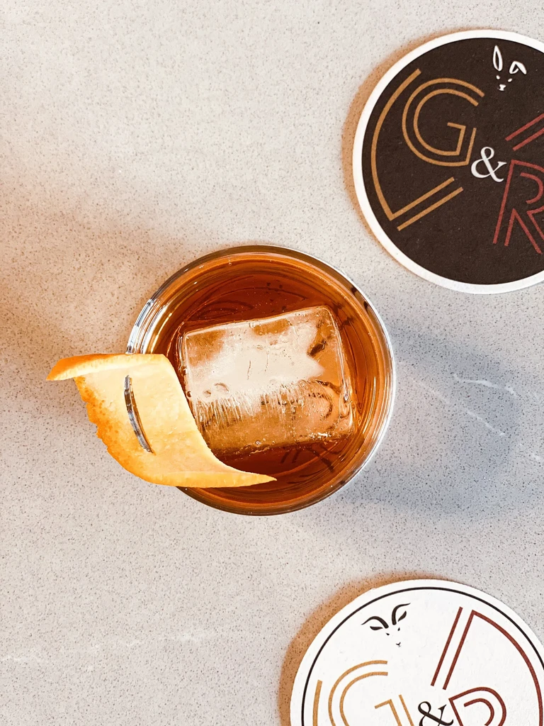 Amber cocktail with square ice, citrus zest, on a grey speckled surface.