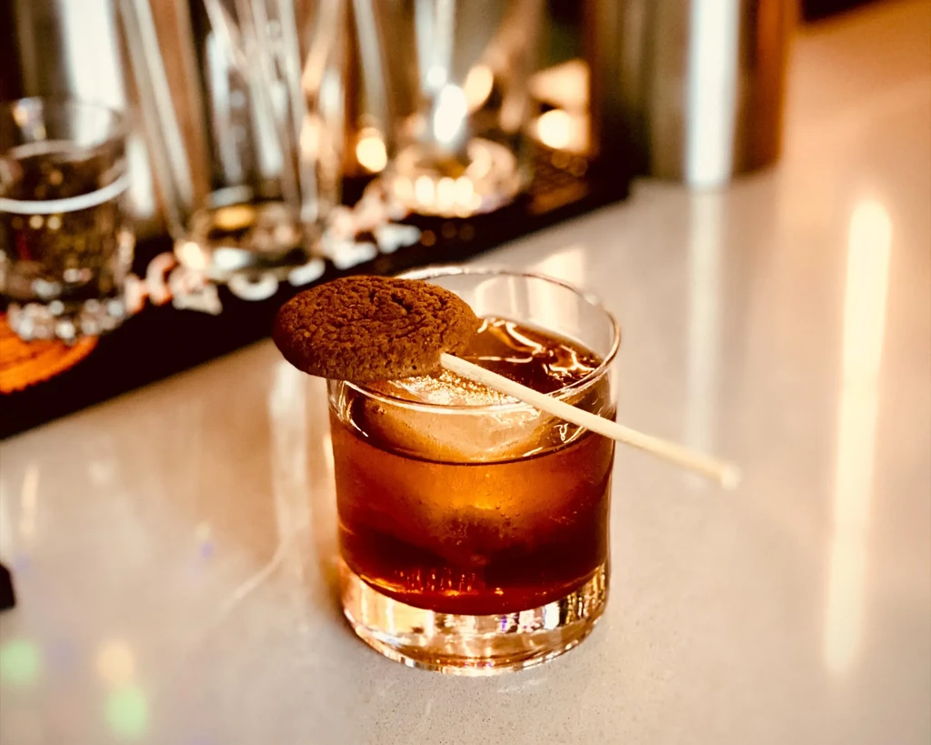 Amber cocktail with large ice cube and cookie on glass rim.