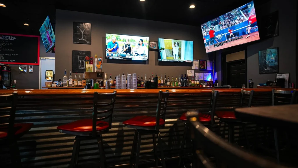 Modern bar interior with long wooden counter, liquor, red stools, and TVs.