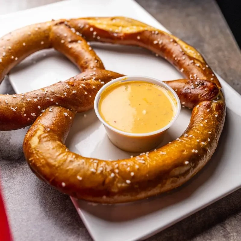 Golden-brown salted pretzel with cheese dip in cup on plate.