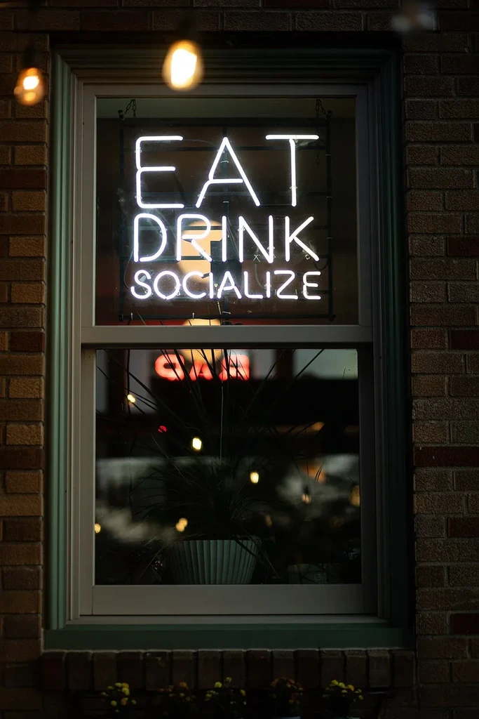 White neon sign "EAT DRINK SOCIALIZE" illuminates a dark building window.