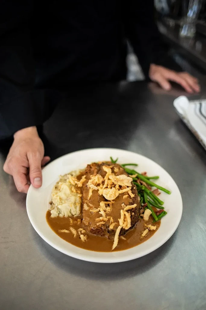 Plated meatloaf, mashed potatoes, green beans, held by uniformed hands.