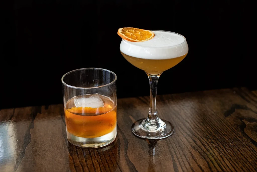 Two cocktails, one iced, one frothy, on dark wood, black background.