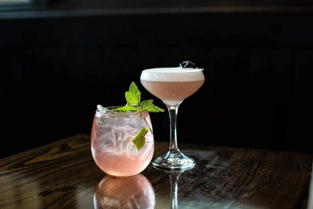 Two pink cocktails with mint and foam on a dark, reflective table.