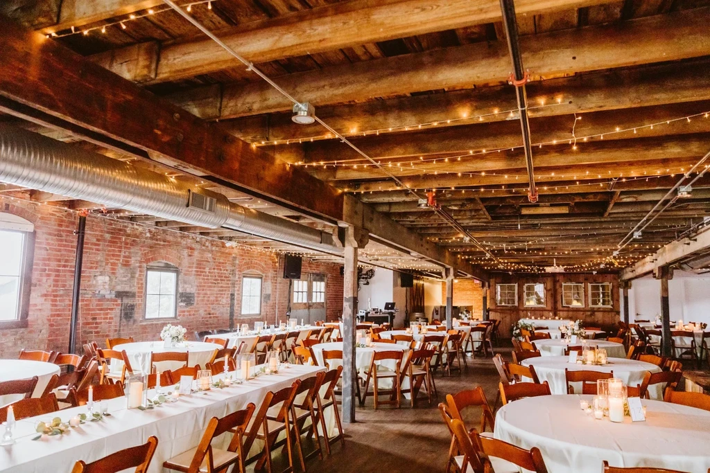 Rustic-industrial event space with tables, chairs, and warm string lights.