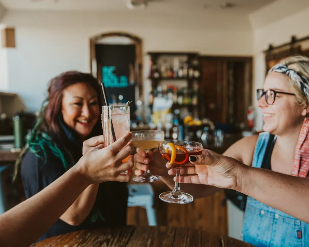 Close-up of three hands toasting colorful cocktails at a bar.