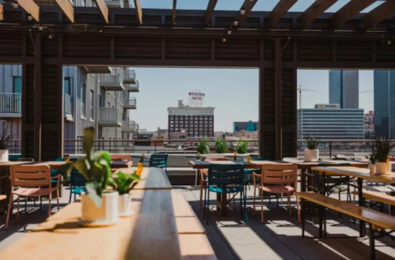 Sunny rooftop dining area with wooden furniture, pergola, city skyline.
