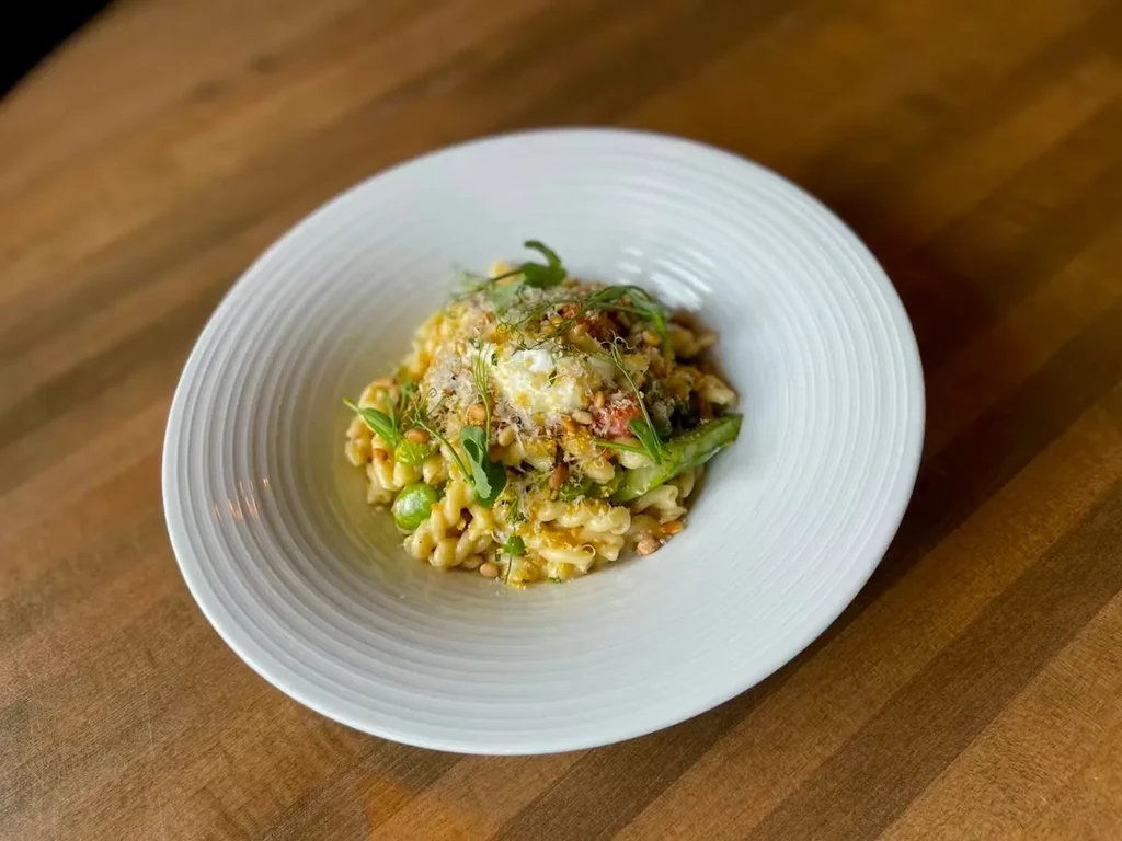 Pasta dish with vegetables, pine nuts, cheese, and creamy topping in bowl.