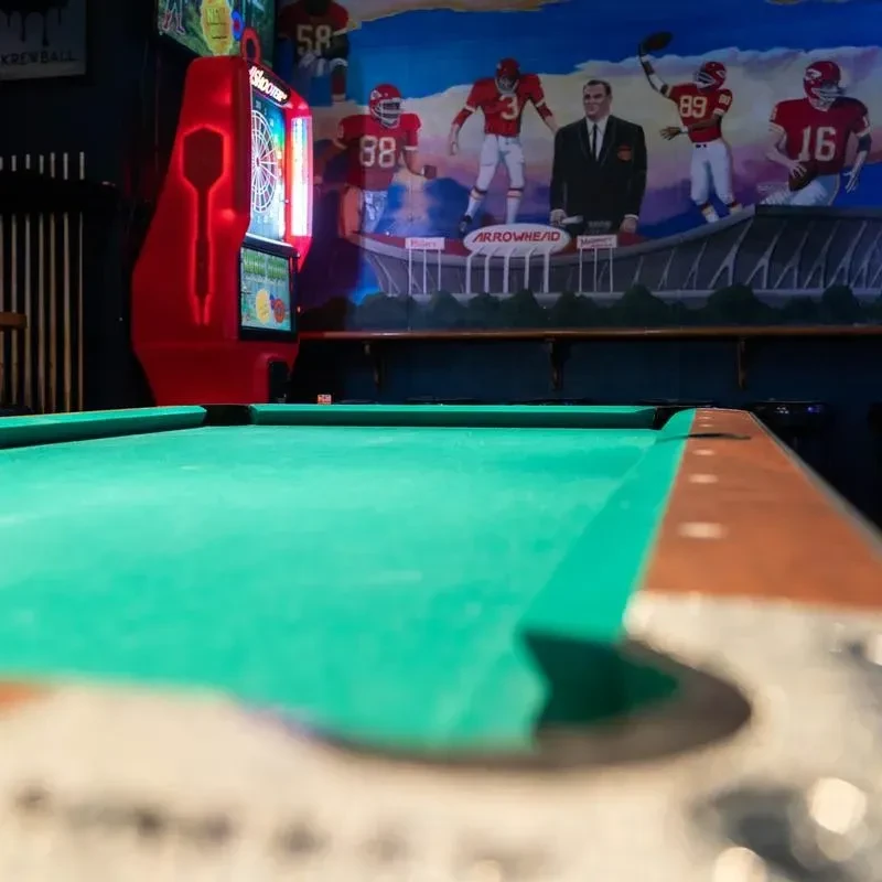 Dim bar with pool table, dartboard, and Arrowhead football mural.