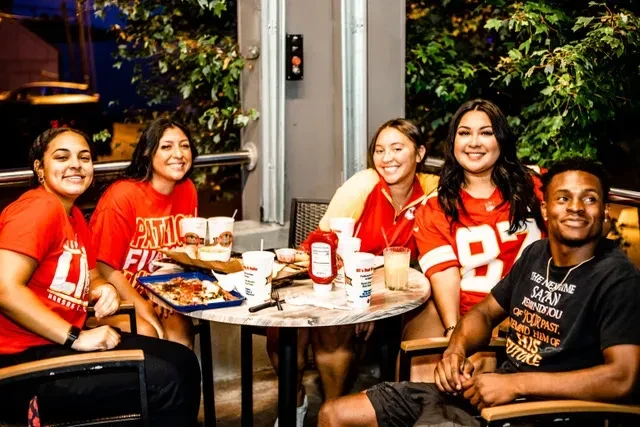 Five smiling young adults at an outdoor restaurant table with food.