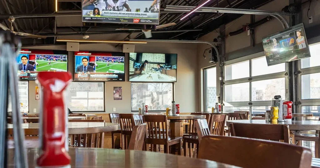 Empty sports bar with wooden tables, large windows, and TVs showing games.