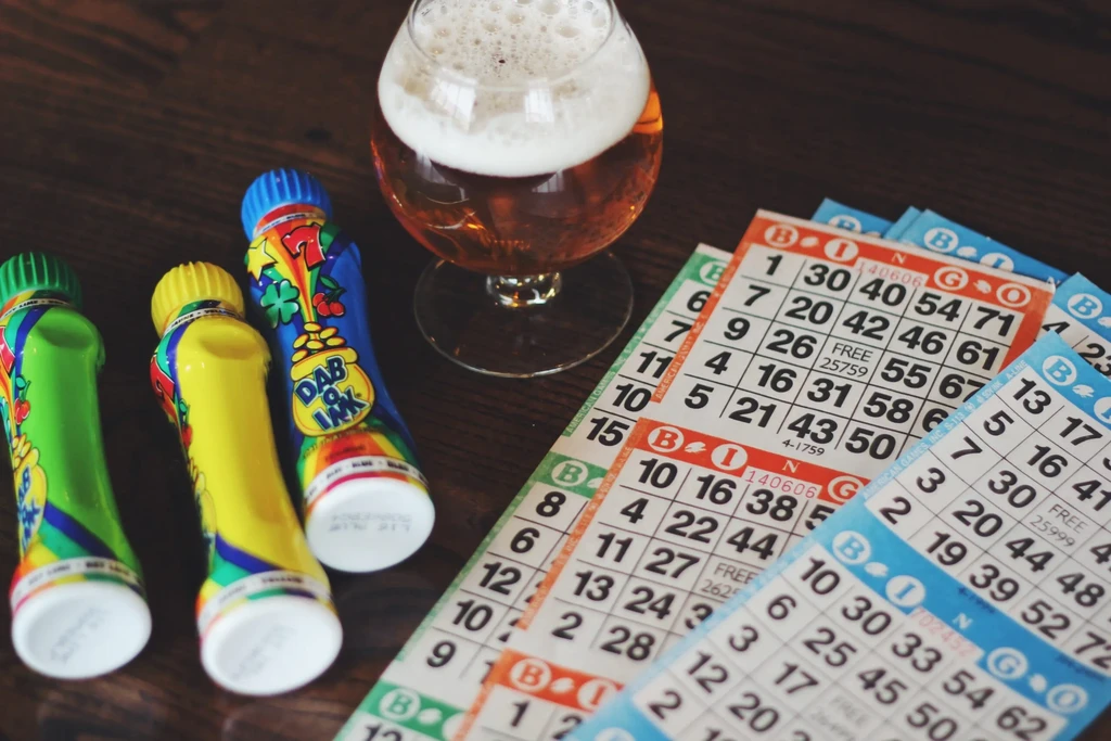 Glass of beer, bingo cards, and daubers on a wooden table.