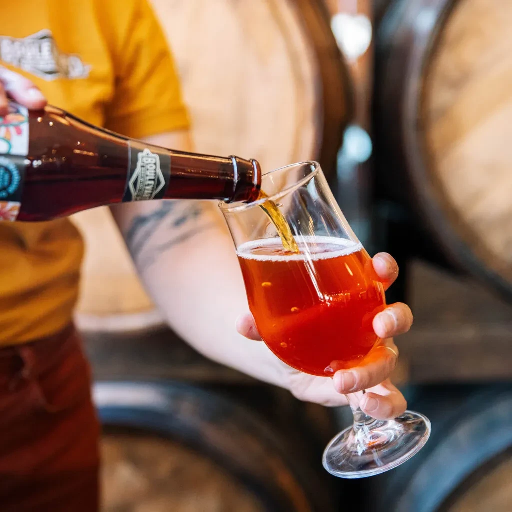 Person pouring Boulevard beer from a bottle, wooden barrels in background.