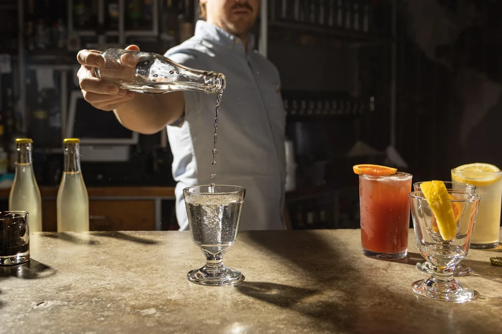Bartender pours drink, droplets falling into glass, other drinks on bar.