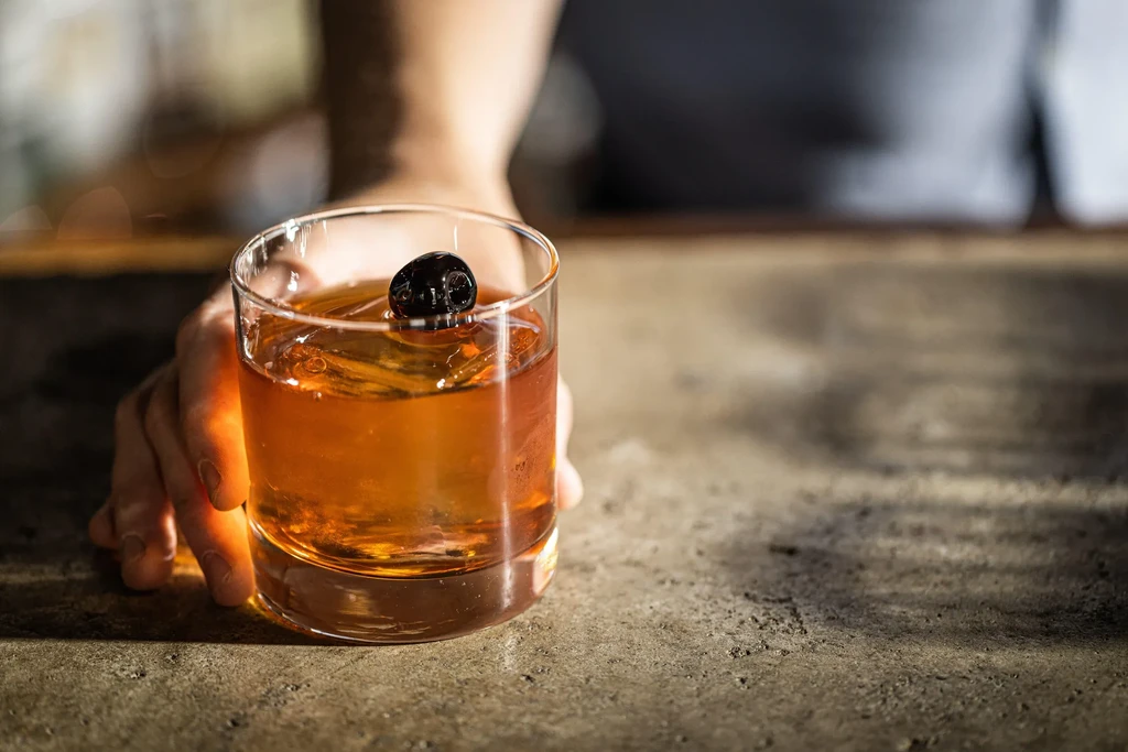 Hand holds amber cocktail with cherry, large ice on stone counter.
