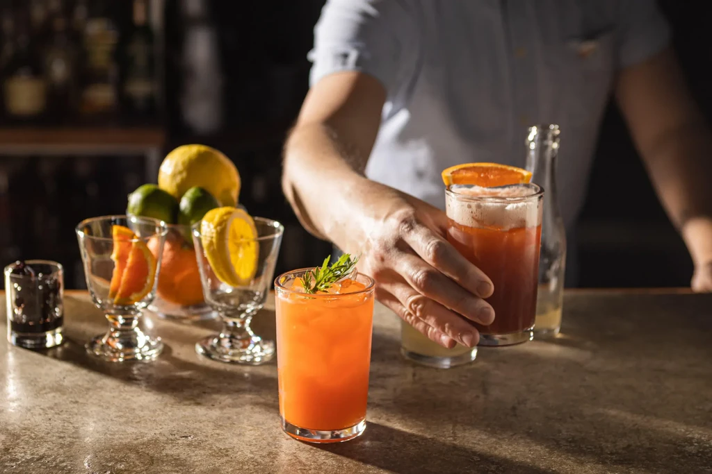 Bar counter with colorful cocktails, fresh citrus, herbs, and hands.