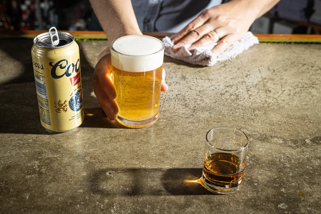 Bartender's hands with beer, spirit glass, Coors can, wiping bar.