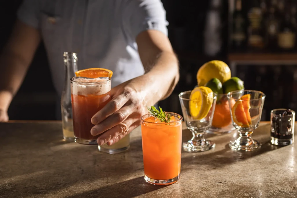 Colorful drinks and garnishes on a bar counter, with a bartender's hand.