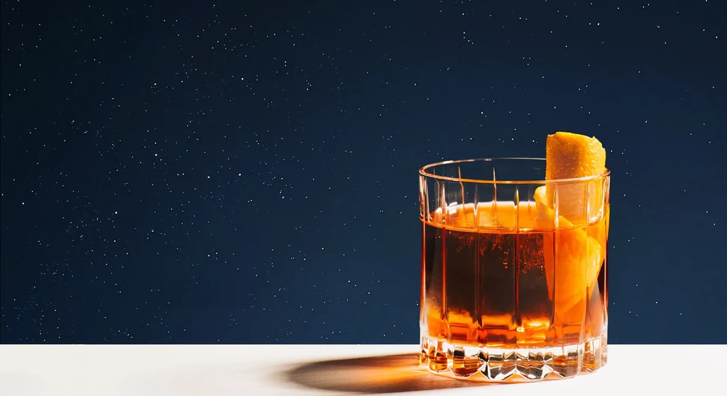 Amber cocktail in faceted glass with orange twist on cosmic background.