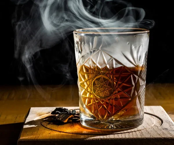Smoked amber cocktail in an intricate cut glass on a wooden coaster.