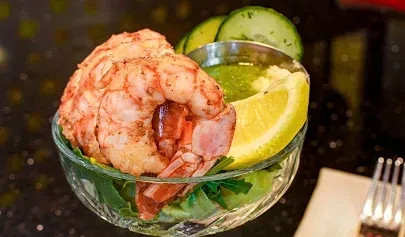 Cooked shrimp cocktail in ornate bowl with lemon, greens, and sauce.