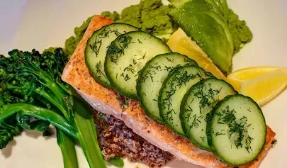 Plated salmon, cucumber, dill, quinoa, broccolini, and lemon wedge.