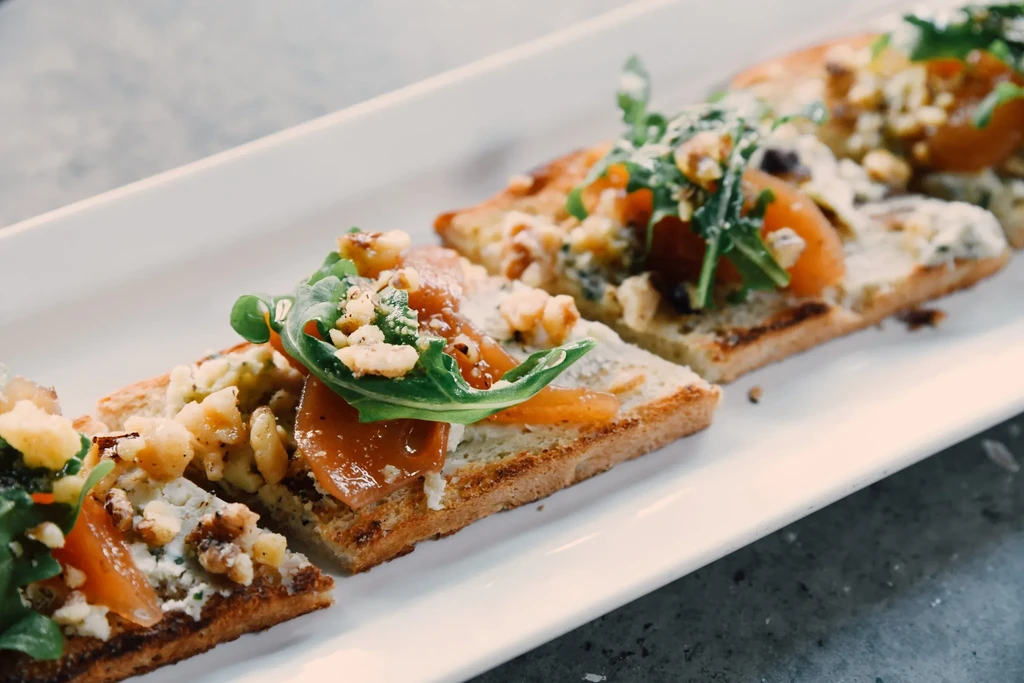 Bruschetta with creamy base, chopped nuts, orange slices, arugula on white plate.
