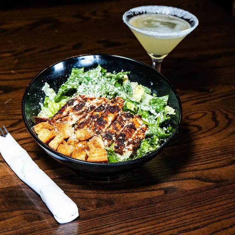 Caesar salad with grilled chicken and a rimmed cocktail on a wooden table.