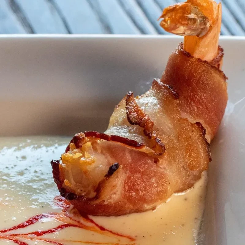 A single bacon-wrapped shrimp with creamy sauce and red drizzle.