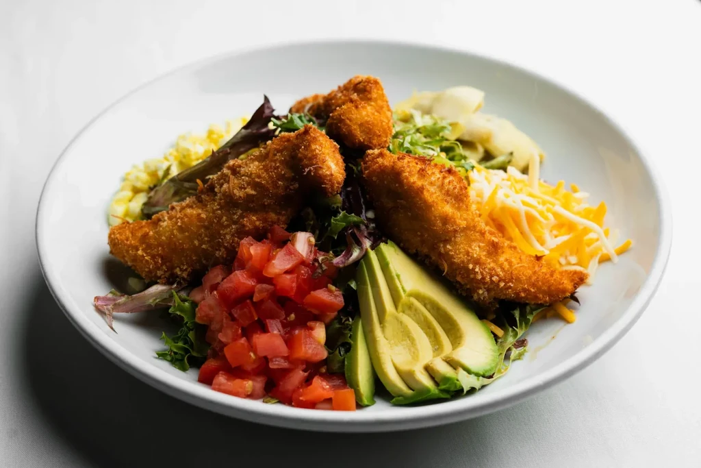 Salad in white bowl: breaded tenders, tomatoes, avocado, cheese, greens.