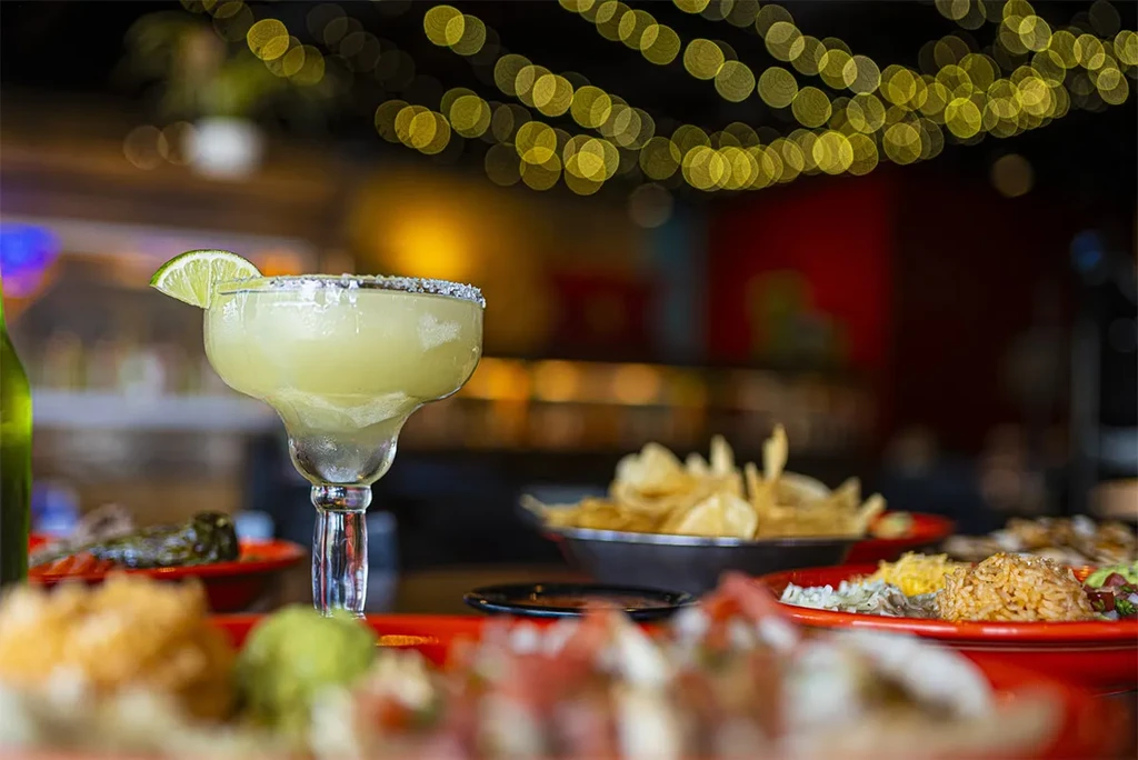 Salt-rimmed lime margarita with lime wedge and blurred restaurant dishes.