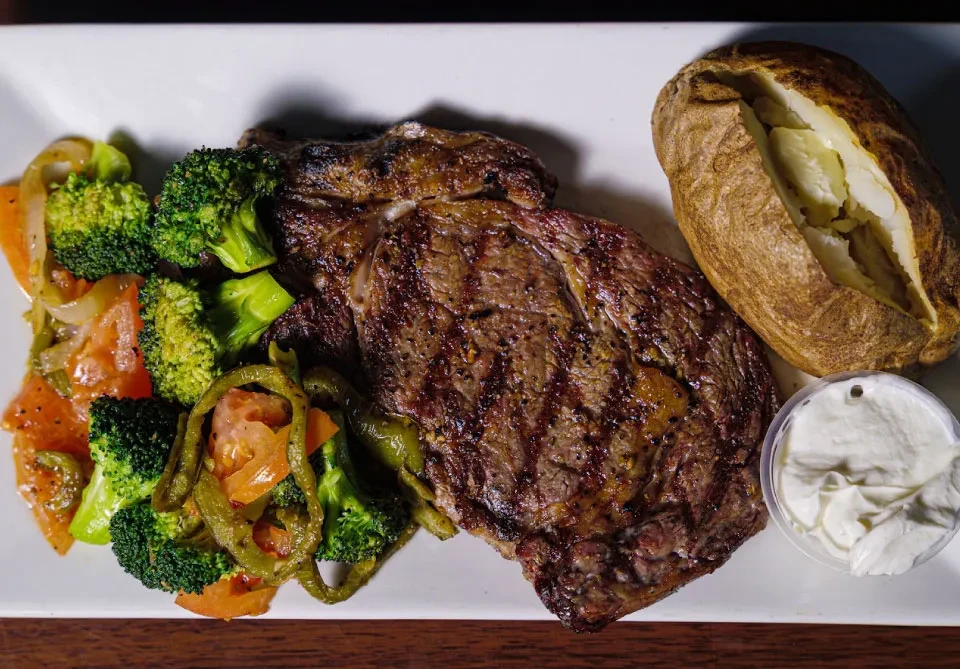 Grilled steak, colorful vegetables, baked potato, and cream on white plate.