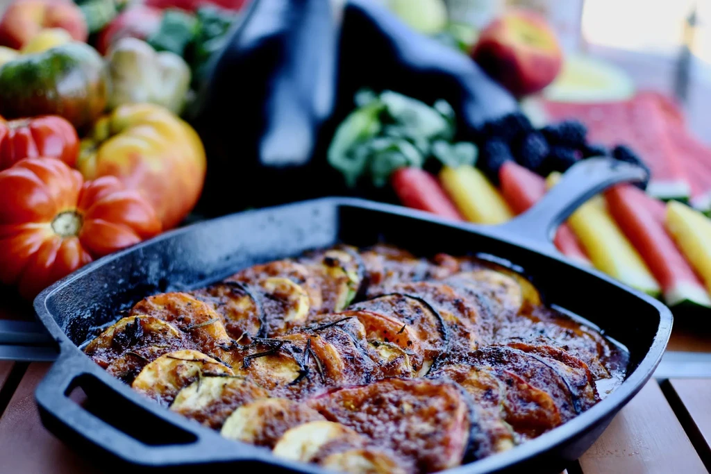 Baked tomato and zucchini dish with rosemary, surrounded by fresh produce.