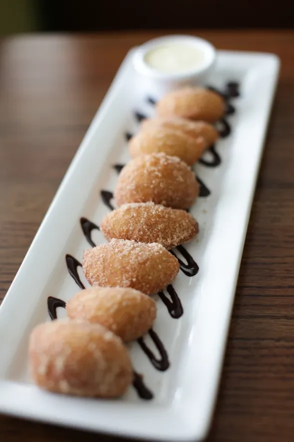 Eight sugar-coated fried dough pieces with chocolate drizzle and dipping sauce.