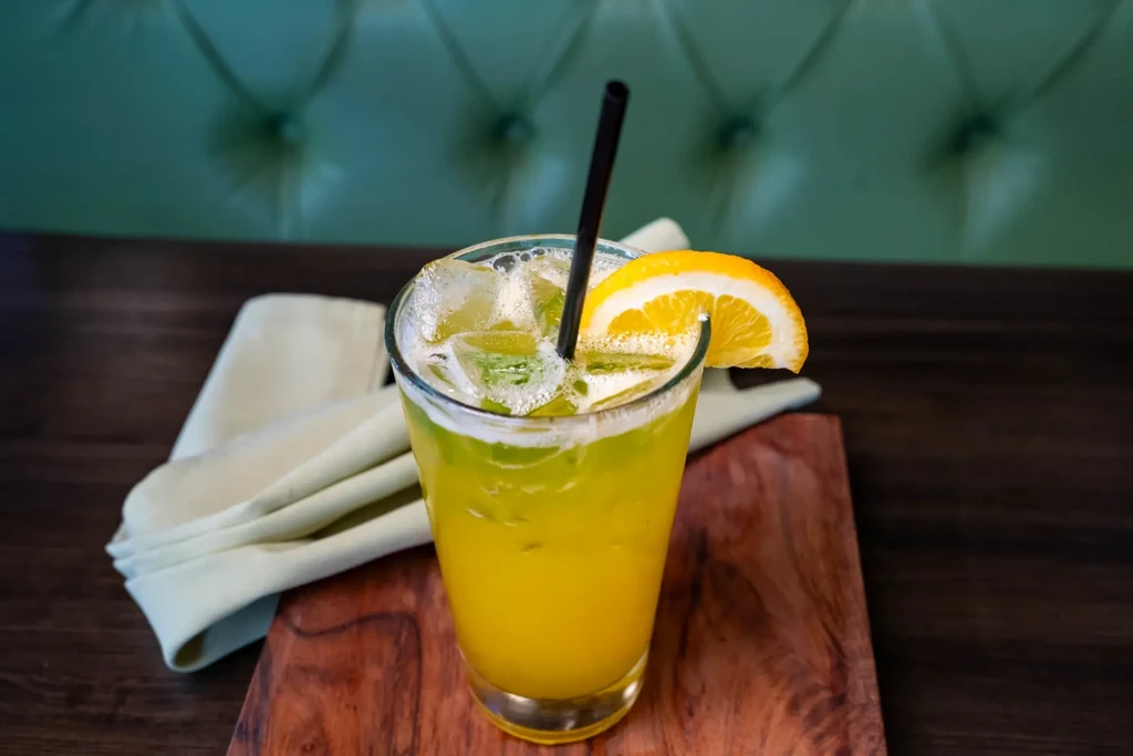 Yellow-green iced drink with orange slice and straw on a wooden board.