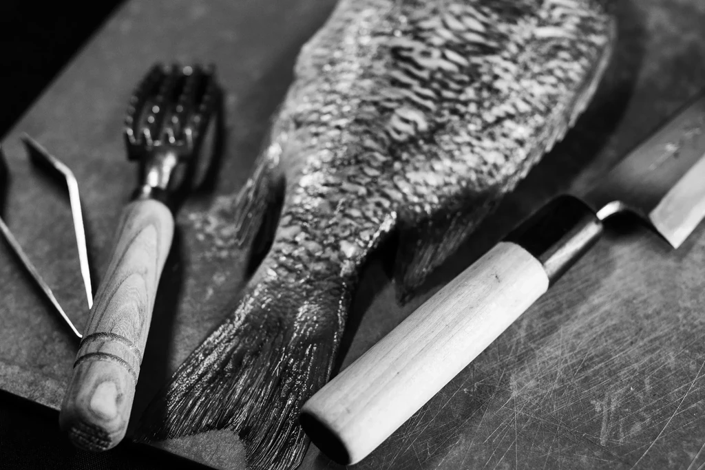 Unscaled raw fish with scaler, tweezers, knife on cutting board.