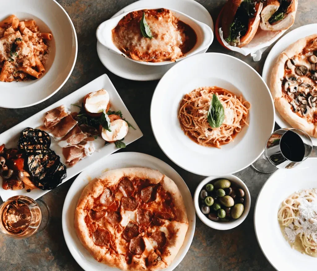 Overhead shot: various Italian dishes, pizzas, and wines on a dark table.