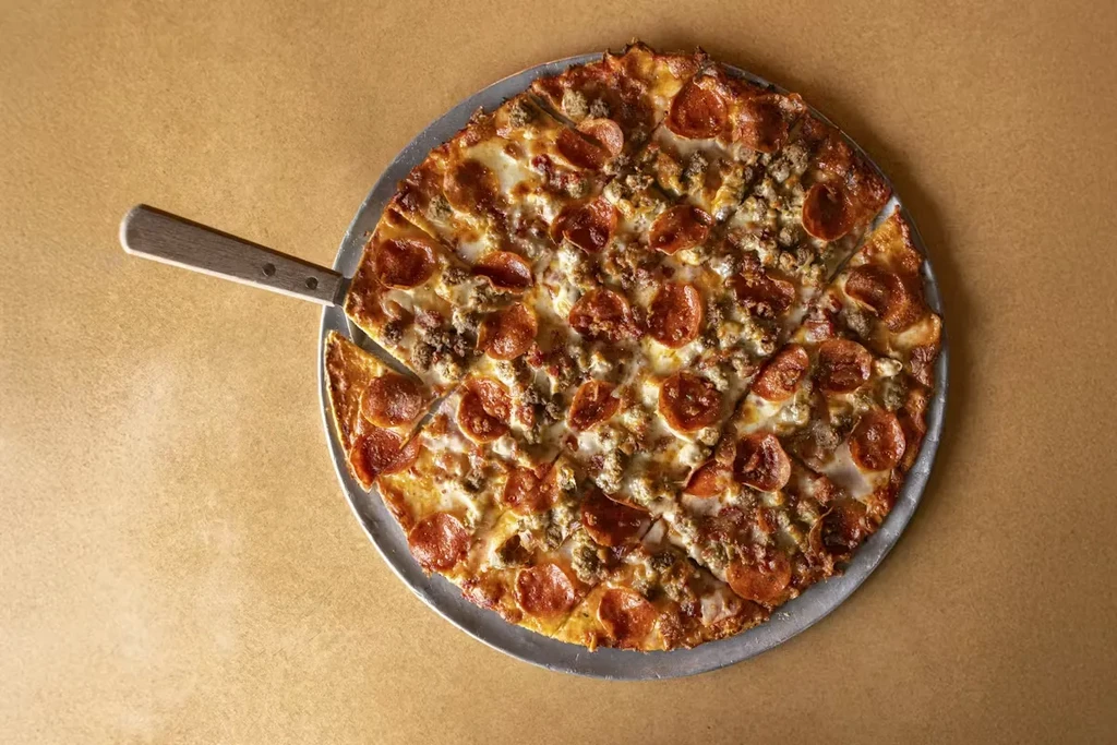 Sliced pizza with cheese, pepperoni, and sausage on pan with server.
