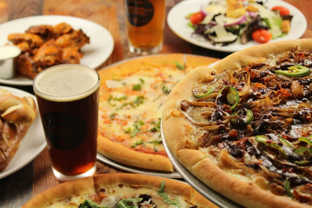 Rustic wooden table spread with pizzas, beer, wings, and salad.